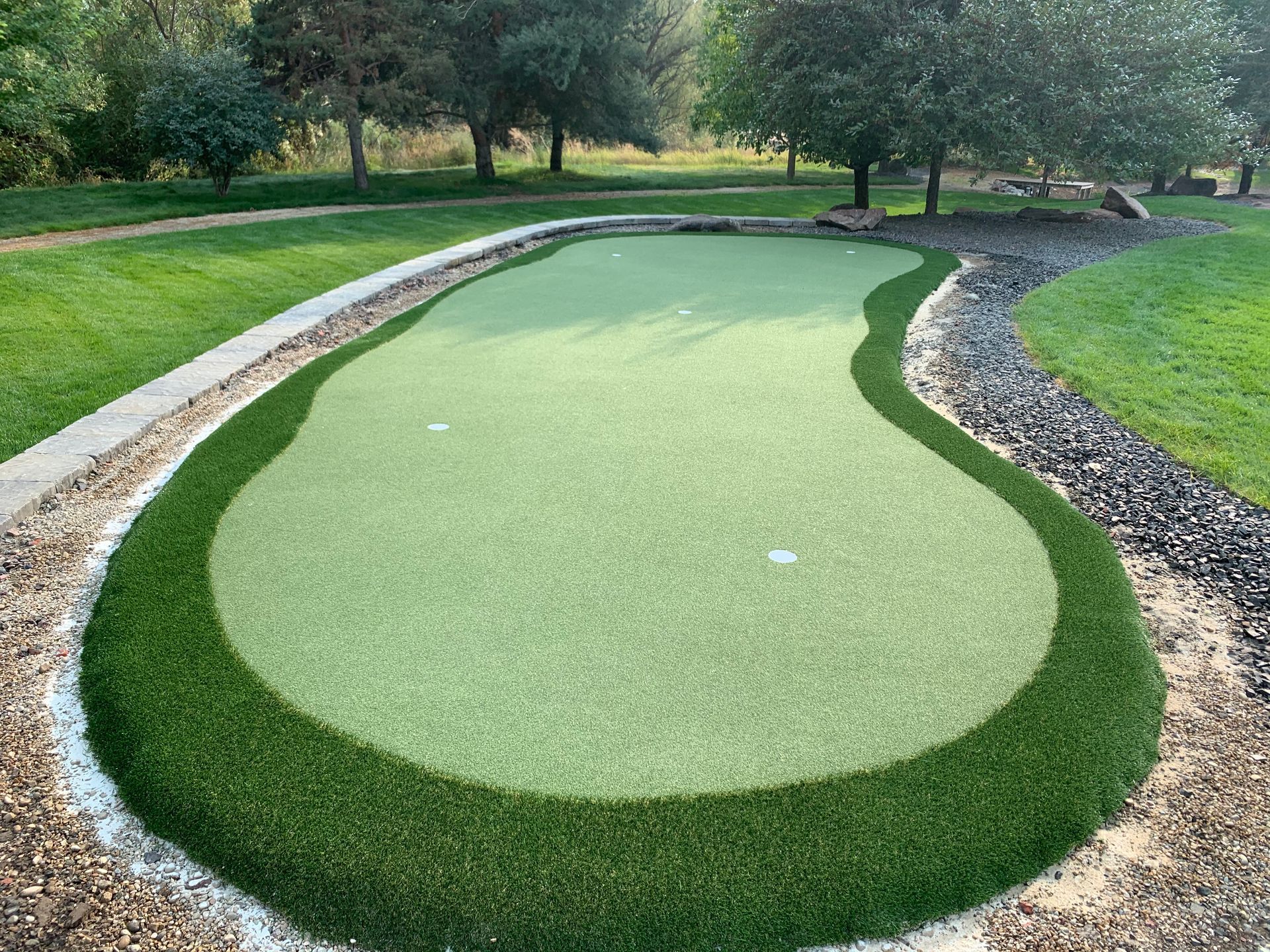 A putting green with a dark green border and light green interior, surrounded by grass and landscaping rocks.