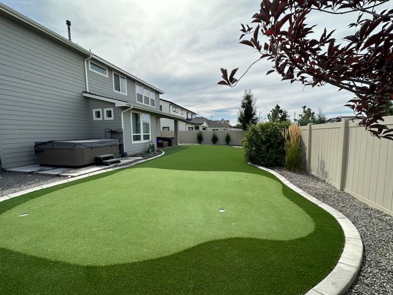 Backyard with a putting green, hot tub, gray house, and a beige fence under a cloudy sky.