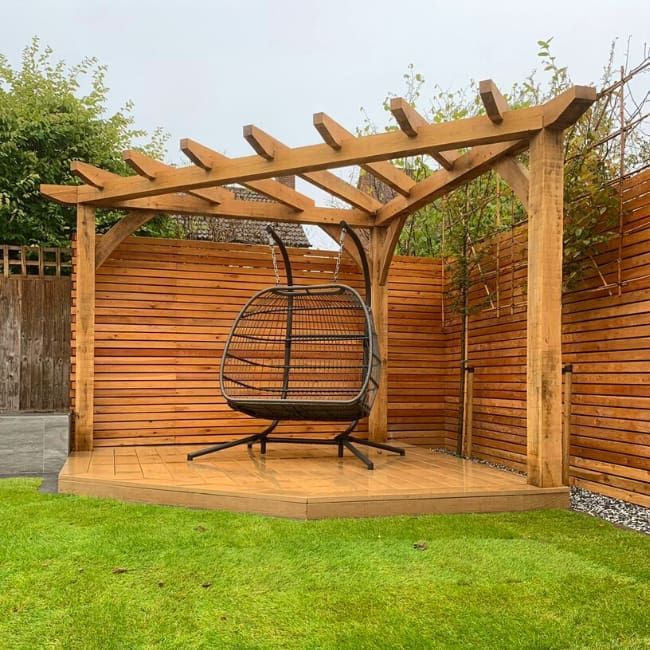 Wooden pergola with hanging chair on a wooden deck in a yard with a wooden fence.