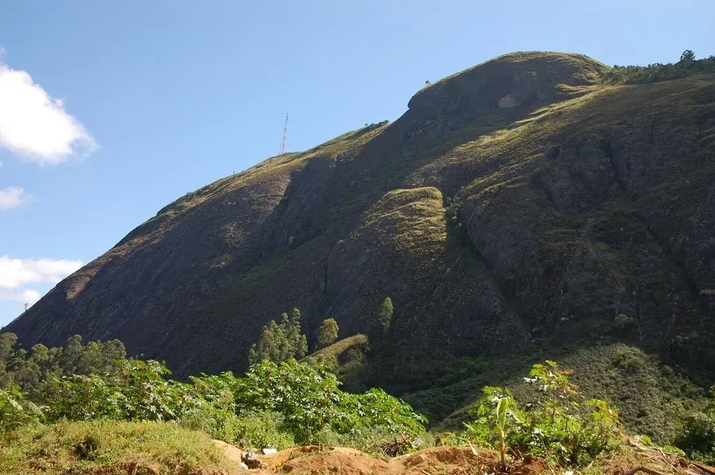 Encosta de montanha ensolarada com vegetação verdejante sob um céu azul e nuvens brancas.