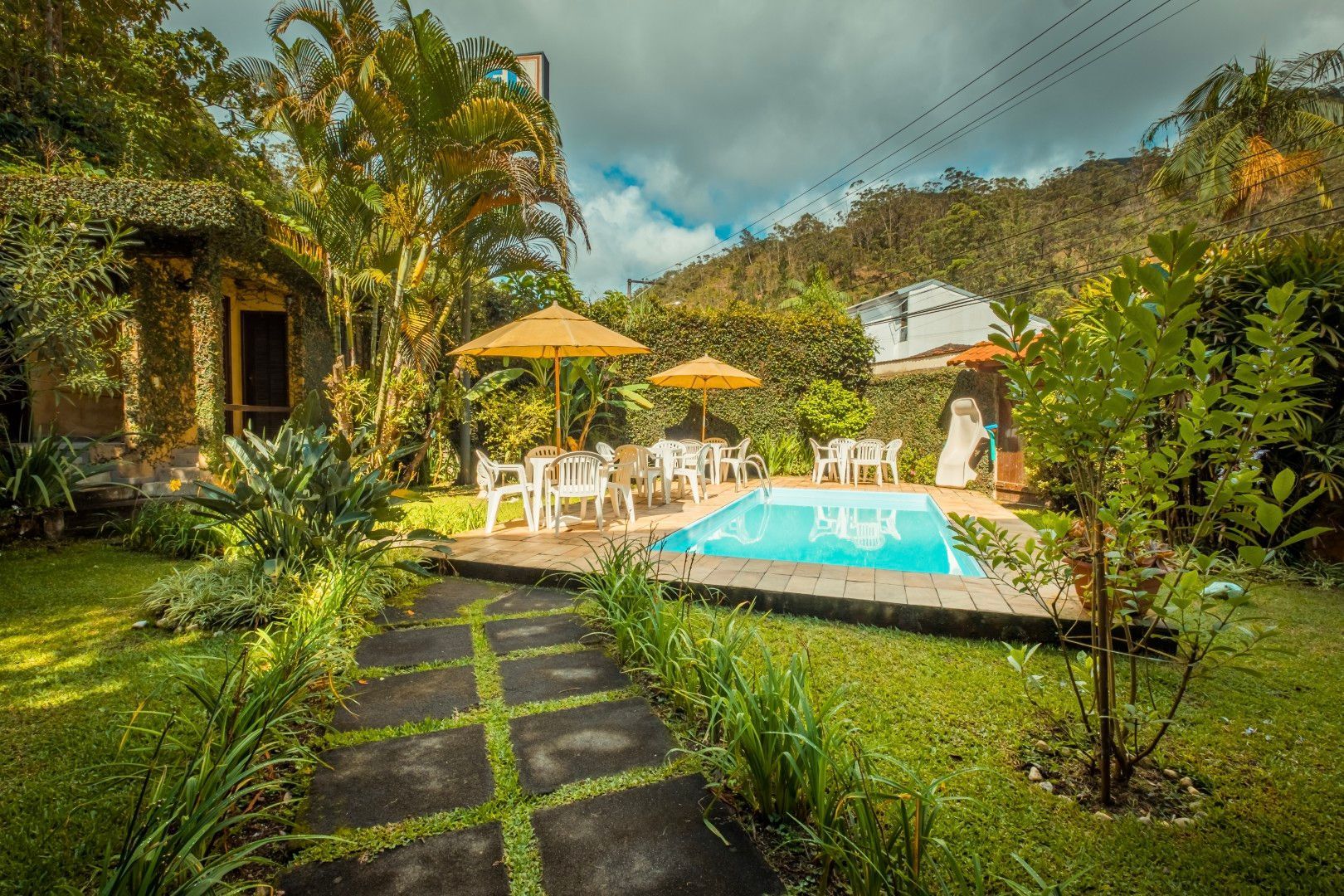Pátio de uma villa tropical com piscina, mesas de jardim, guarda-sóis e vegetação exuberante sob um céu nublado.