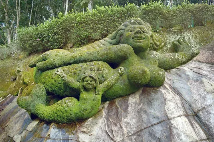 Escultura de pedra reclinada coberta de musgo sobre uma rocha inclinada, com uma figura menor abaixo dela em um ambiente florestal.
