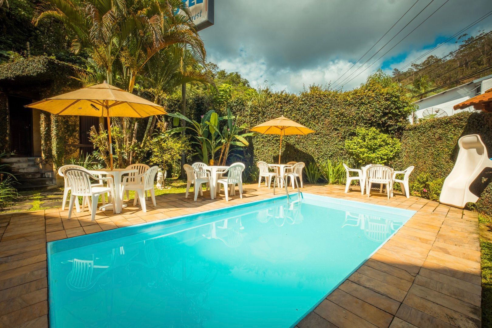 Pátio tropical com piscina, cadeiras brancas, guarda-sóis amarelos e vegetação exuberante sob um céu nublado.