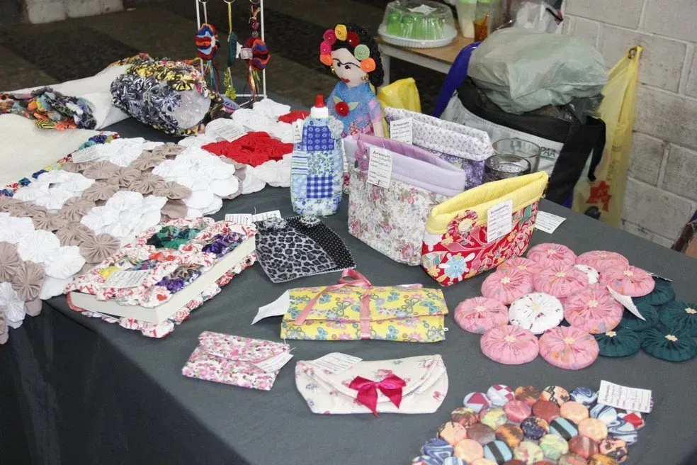 Artesanatos têxteis feitos à mão expostos sobre uma mesa, incluindo cestos, bolsas e arranjos florais em cores vibrantes.