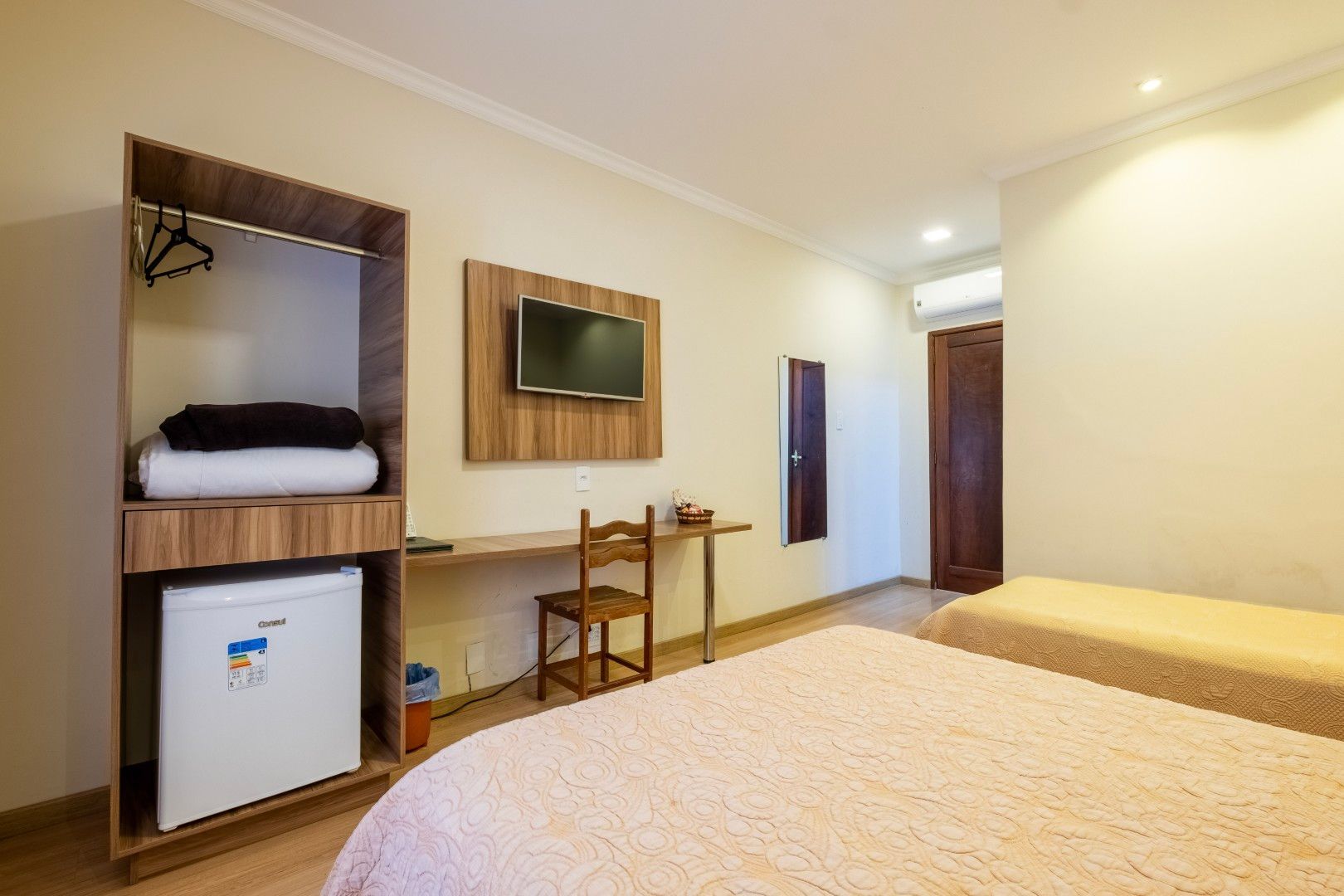 Quarto de hotel pequeno com duas camas, TV de parede, escrivaninha, cadeira e frigobar em um ambiente iluminado.