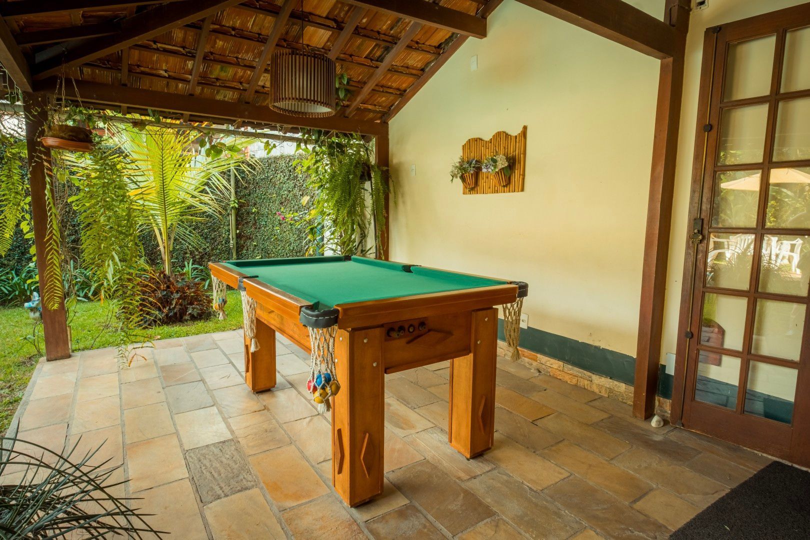Pátio coberto com mesa de bilhar de madeira, piso de azulejo e vista para o jardim aberto.