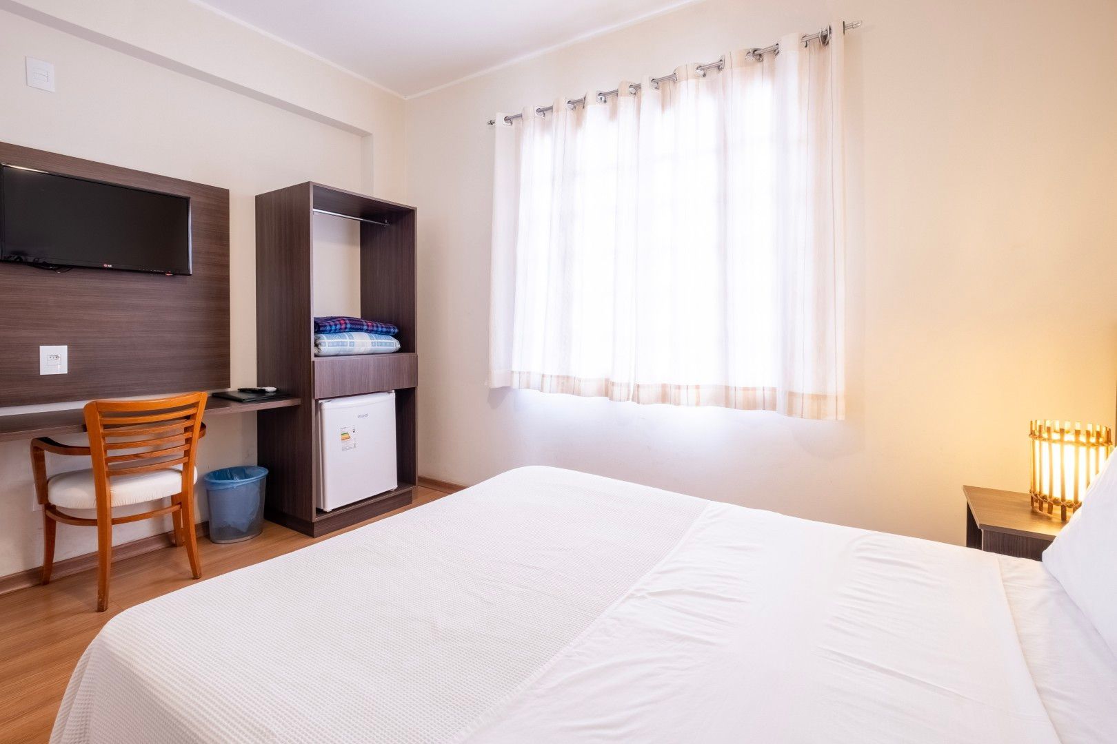 Quarto iluminado com cama, escrivaninha, TV, janela e iluminação aconchegante de abajur.