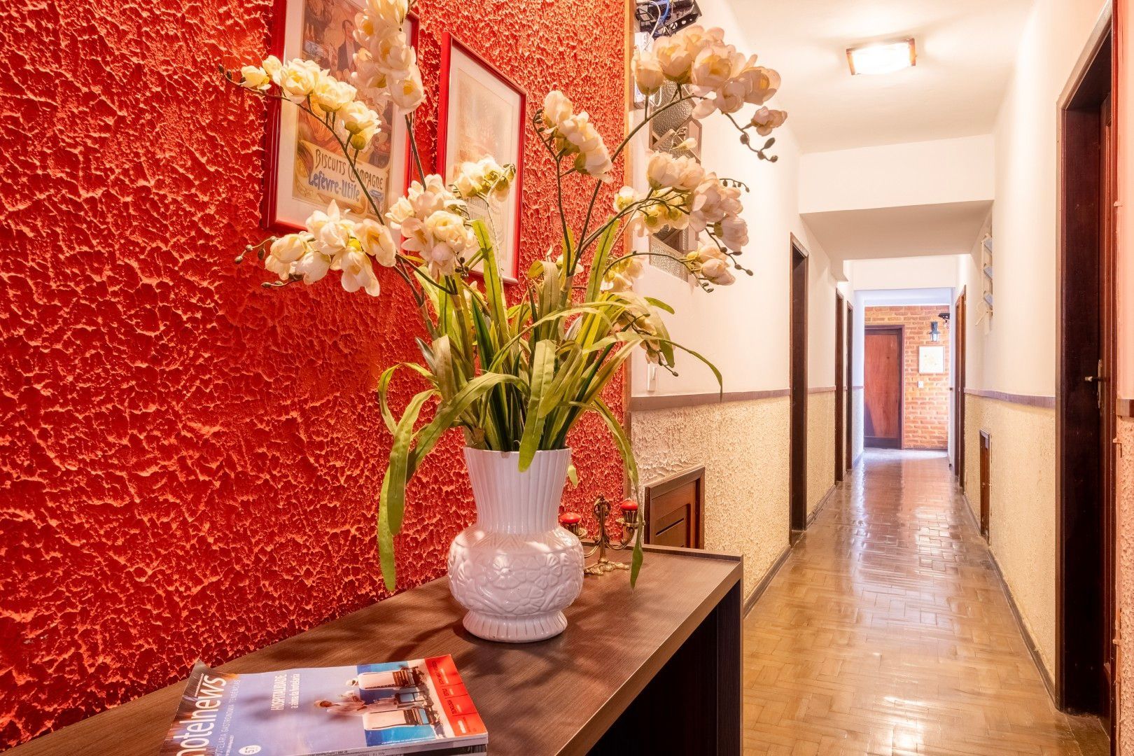 Corredor com parede texturizada em vermelho, mesa de console escura, vaso de flores brancas e um corredor iluminado além.