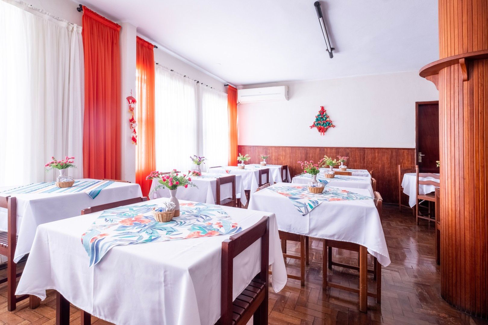 Sala de jantar iluminada com toalhas de mesa brancas, cadeiras de madeira, cortinas laranja e piso de madeira polida.