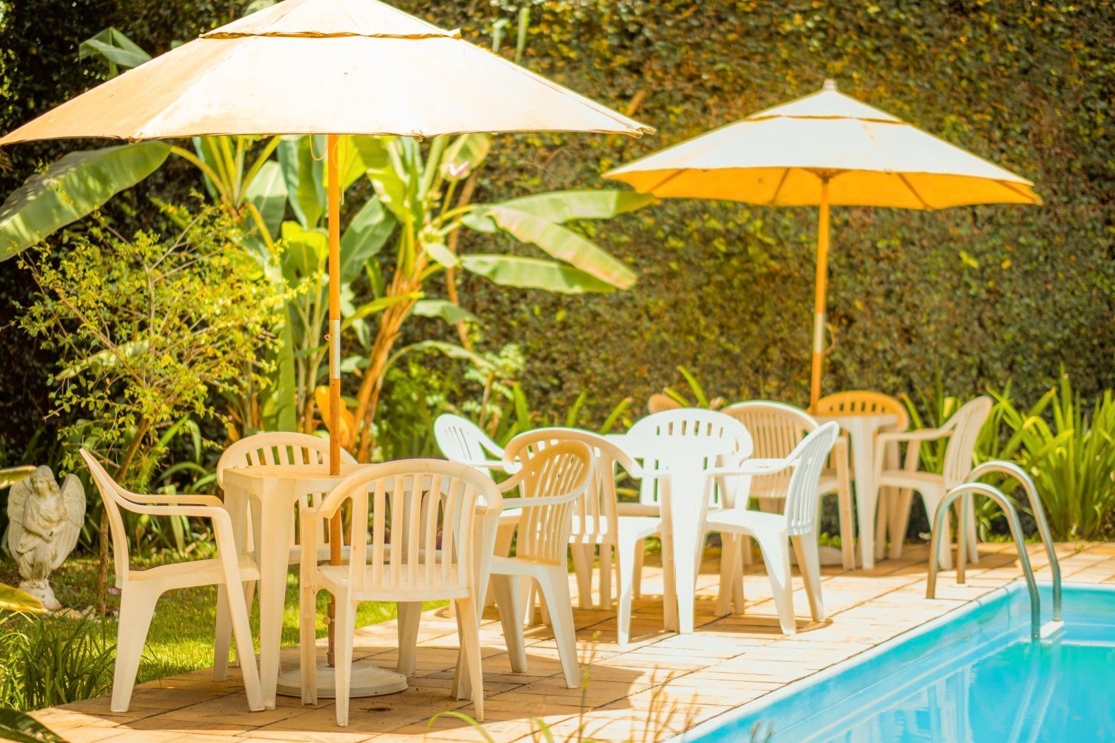 Pátio à beira da piscina com mesas, cadeiras e guarda-sóis brancos ao lado de uma vegetação exuberante e uma piscina azul.