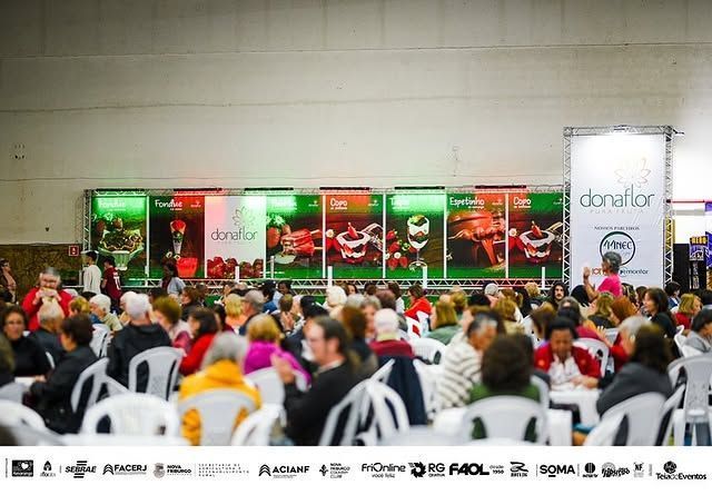 Público sentado em frente a um palco com bandeiras vermelhas e verdes em um evento em local fechado.