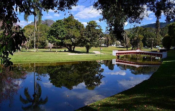 Parque ensolarado com um lago refletor, árvores verdes e uma pequena ponte vermelha sob um céu azul.
