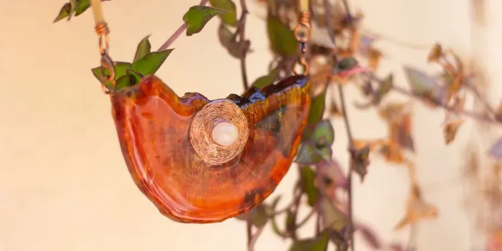 Um vaso suspenso translúcido laranja com uma pequena planta e uma decoração redonda central contra uma parede bege.