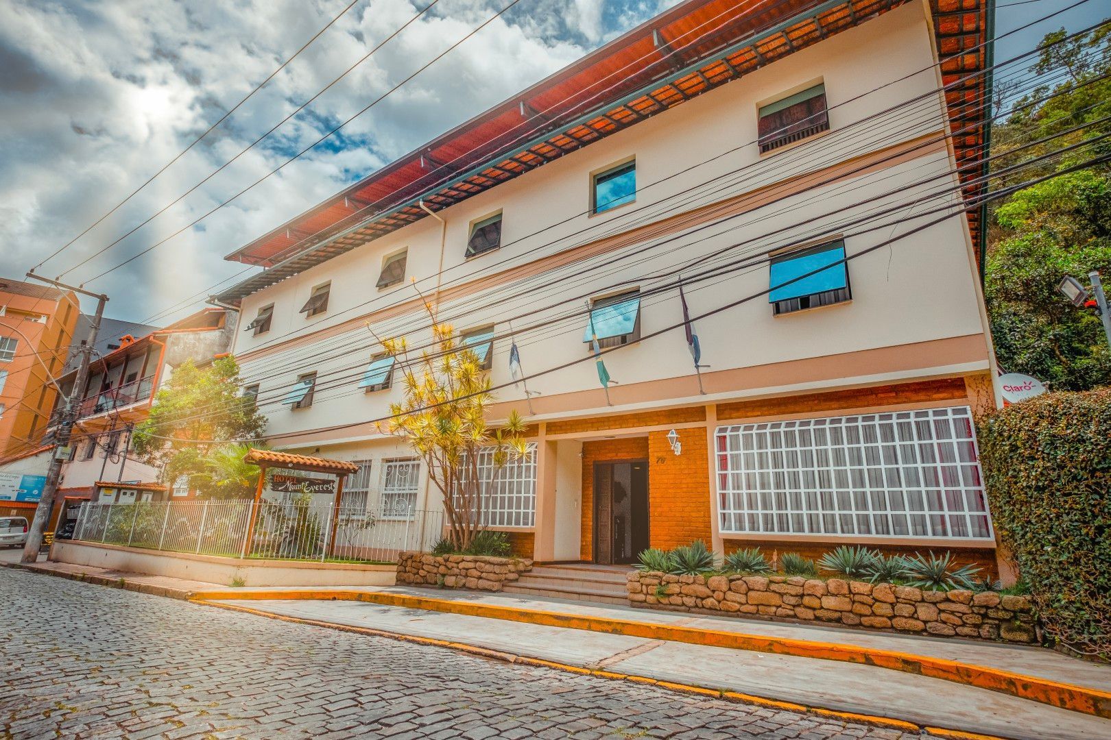 Prédio moderno de vários andares com paredes bege, detalhes laranja no telhado e rua de paralelepípedos em frente.