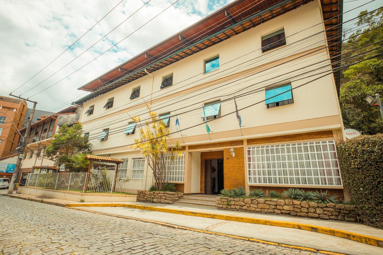 Prédio bege de três andares em rua inclinada, com muro de pedra e entrada para garagem.