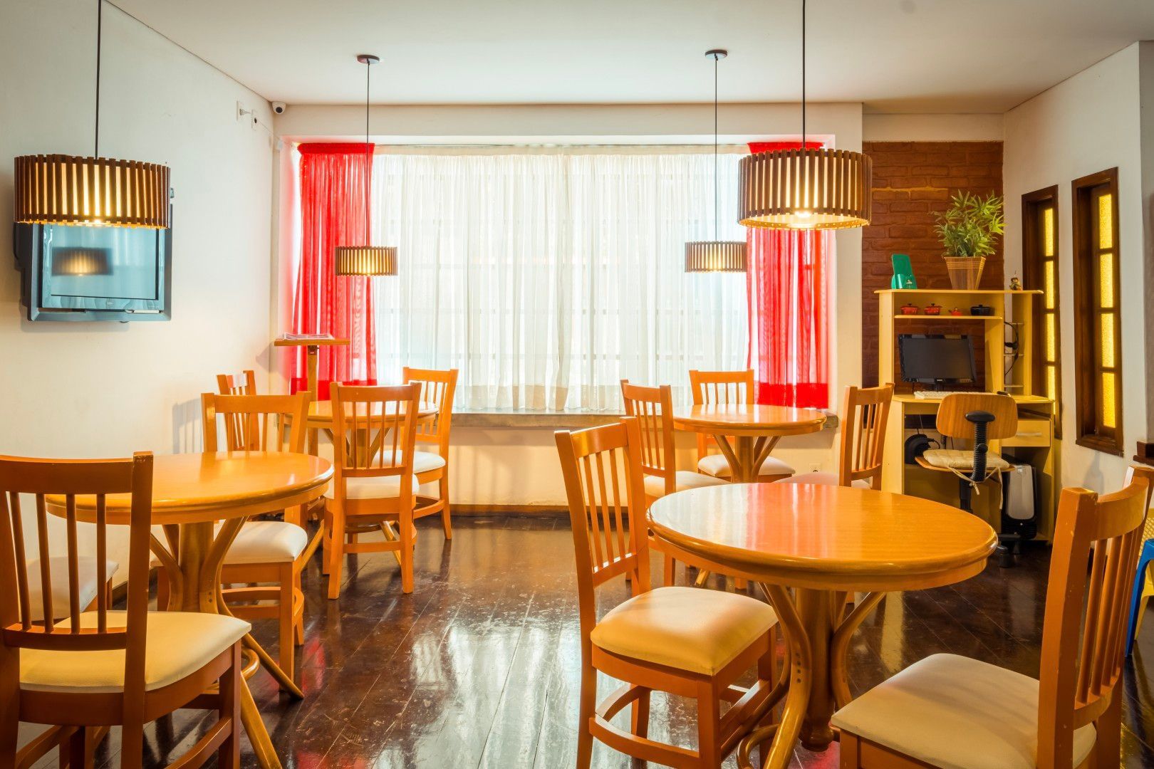 Sala de jantar iluminada com mesas e cadeiras de madeira, janelas amplas e luminárias pendentes em tons quentes.