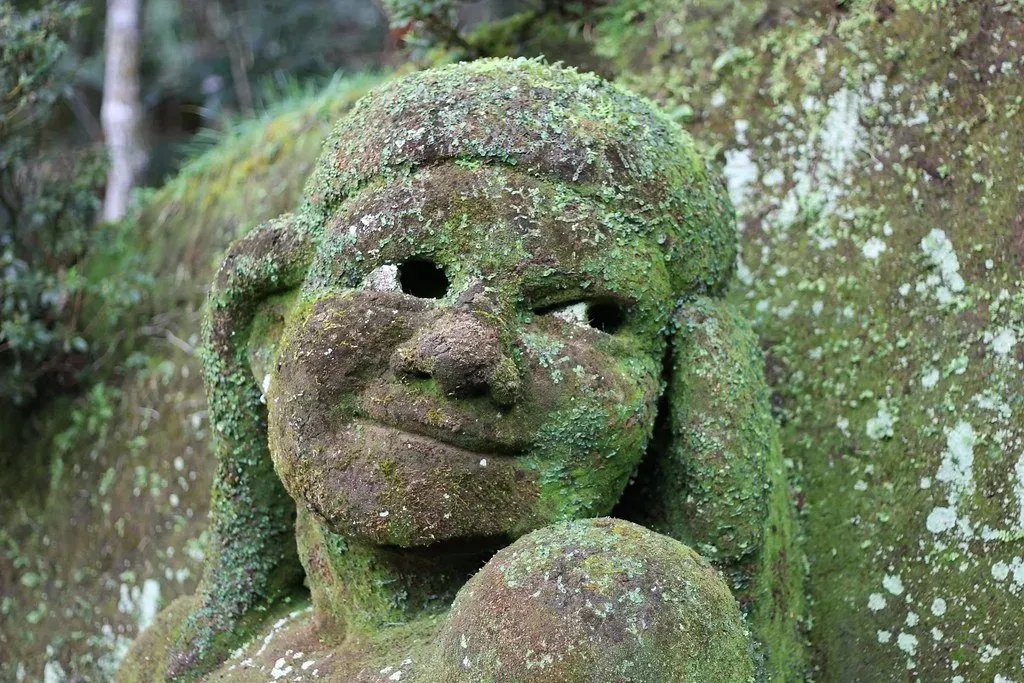 Escultura de pedra coberta de musgo com expressão serena em um ambiente florestal.
