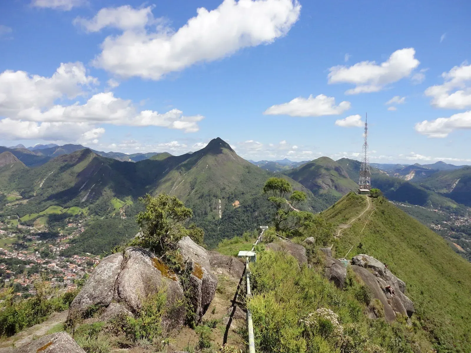 Mirante no topo da montanha com trilha rochosa, picos verdes e céu azul com nuvens.