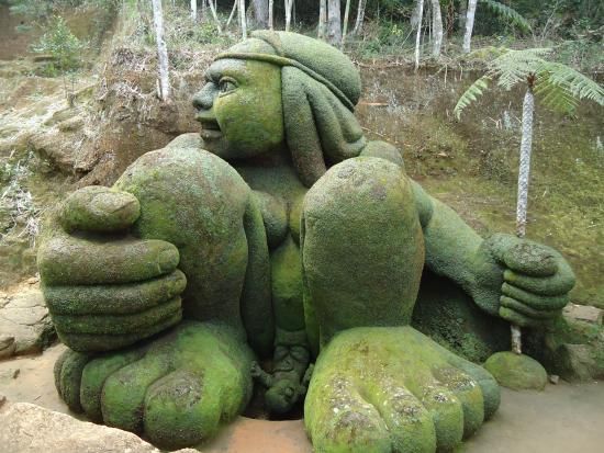 Escultura de pedra coberta de musgo representando um gigante sentado em um ambiente florestal.