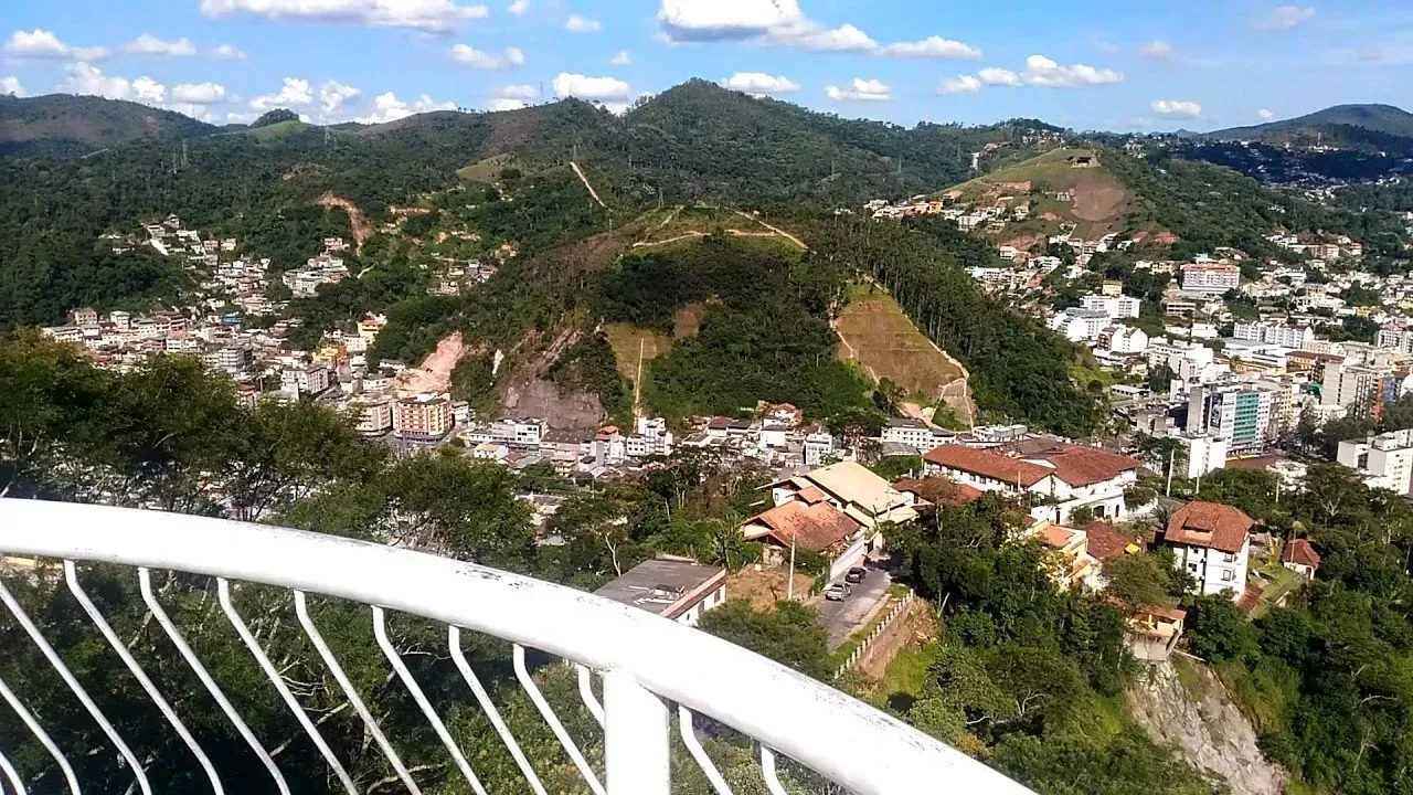 Mirante na montanha com corrimão branco, com vista para uma cidade e colinas verdejantes sob um céu azul.
