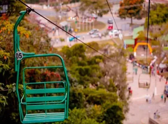 Teleférico turquesa sobre uma passarela desfocada do parque, com árvores e pessoas ao fundo.