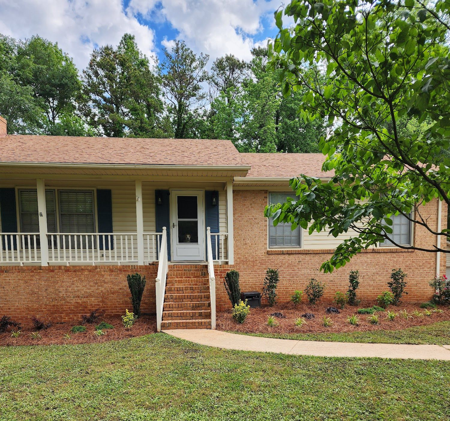 Tan brick ranch-style house with a porch and steps, surrounded by green grass and trees.