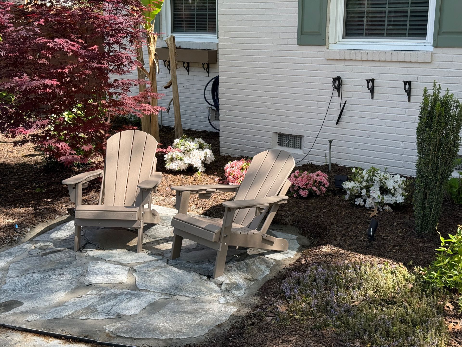 Two Adirondack chairs on a stone patio by a white brick house, surrounded by flowers and plants.