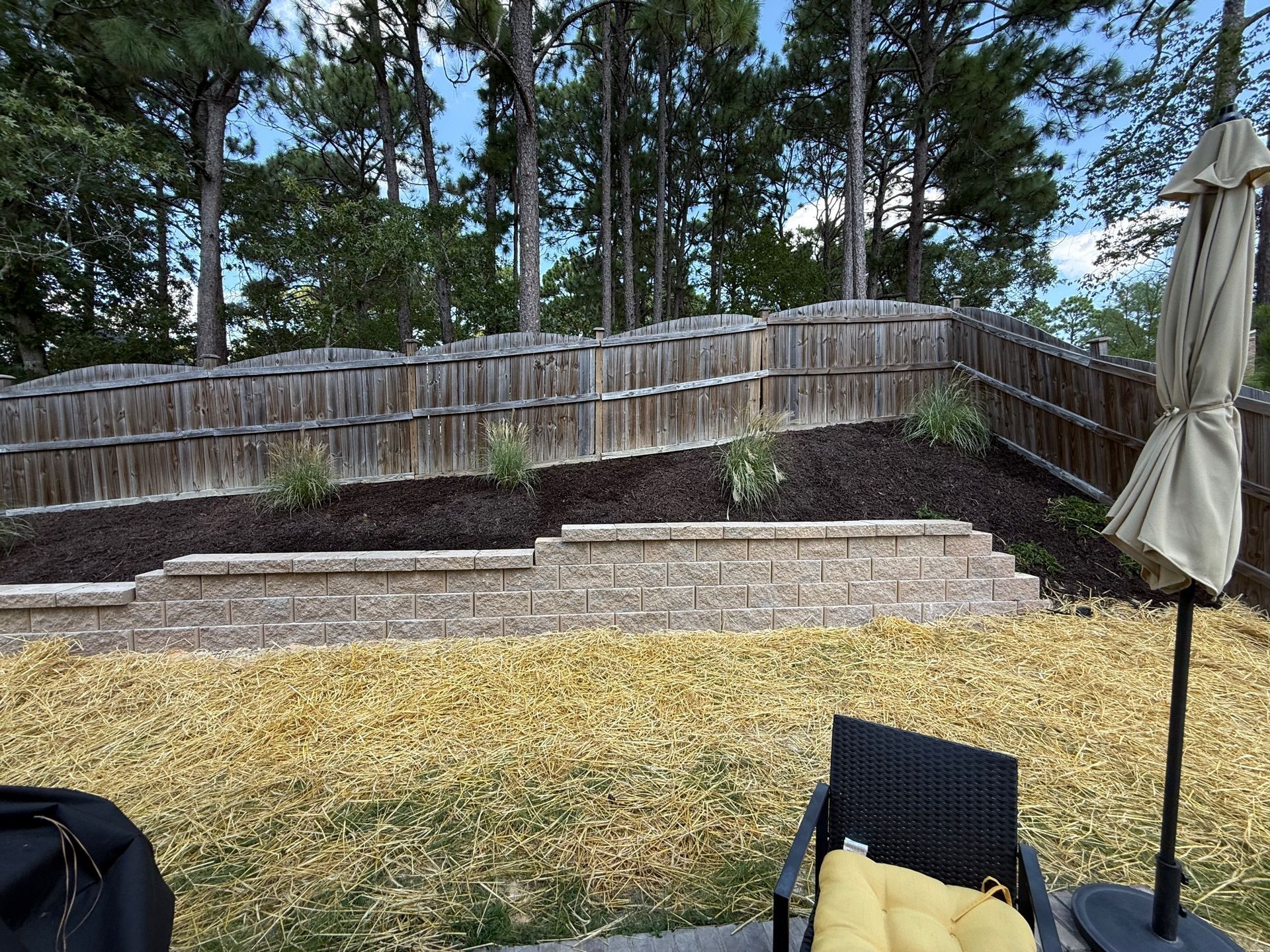 Backyard with a retaining wall, mulch, plants, and a wooden fence. A patio umbrella and chair are in the foreground.