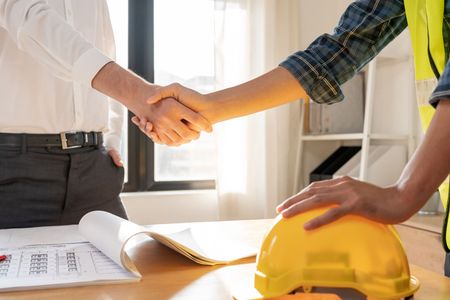 two men are shaking hands over a table with a hard hat on it