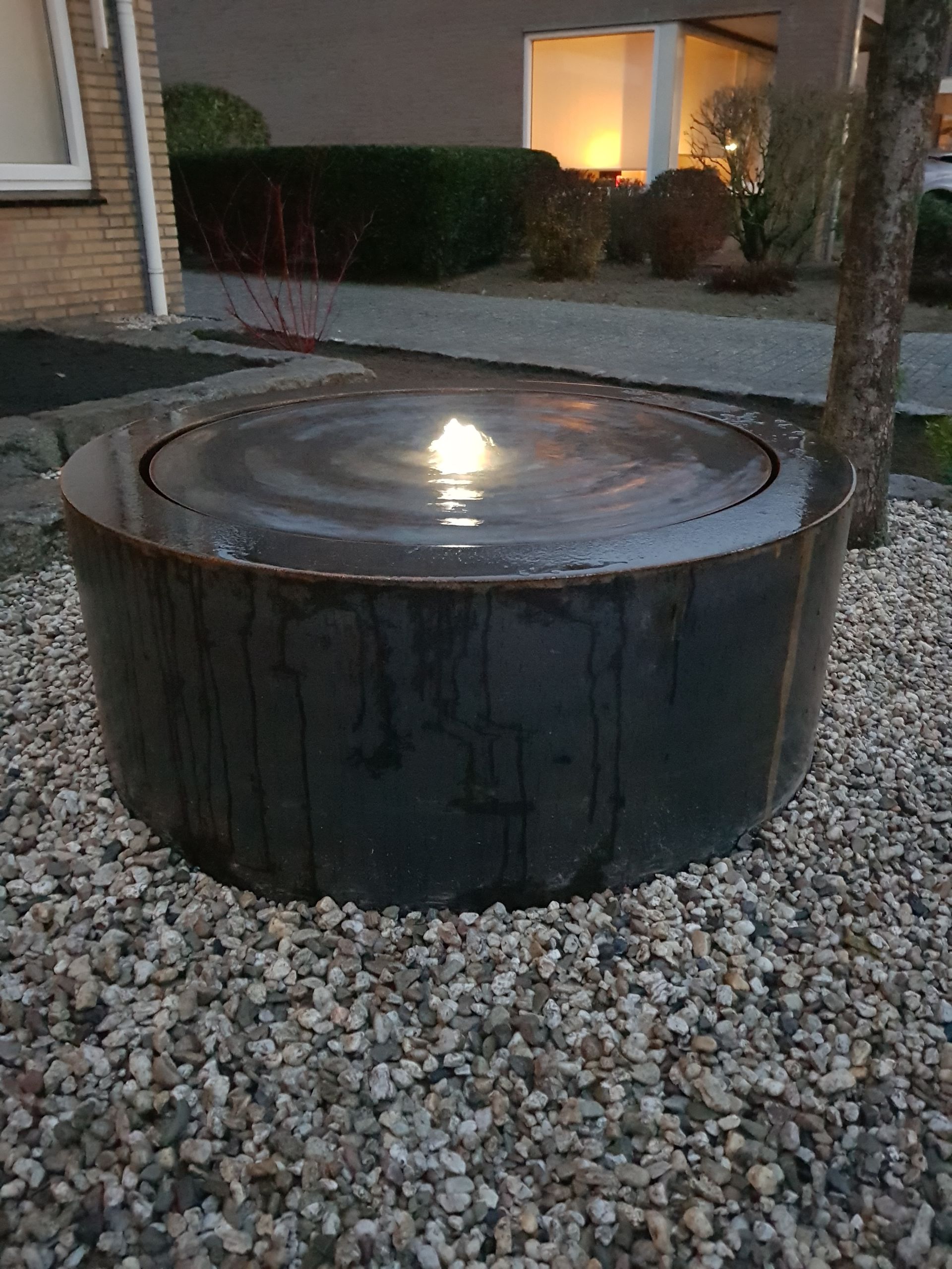 Zwarte, cilindrische waterpartij voor buiten met een verlicht middenstuk, geplaatst in een grindhofje naast een huis.