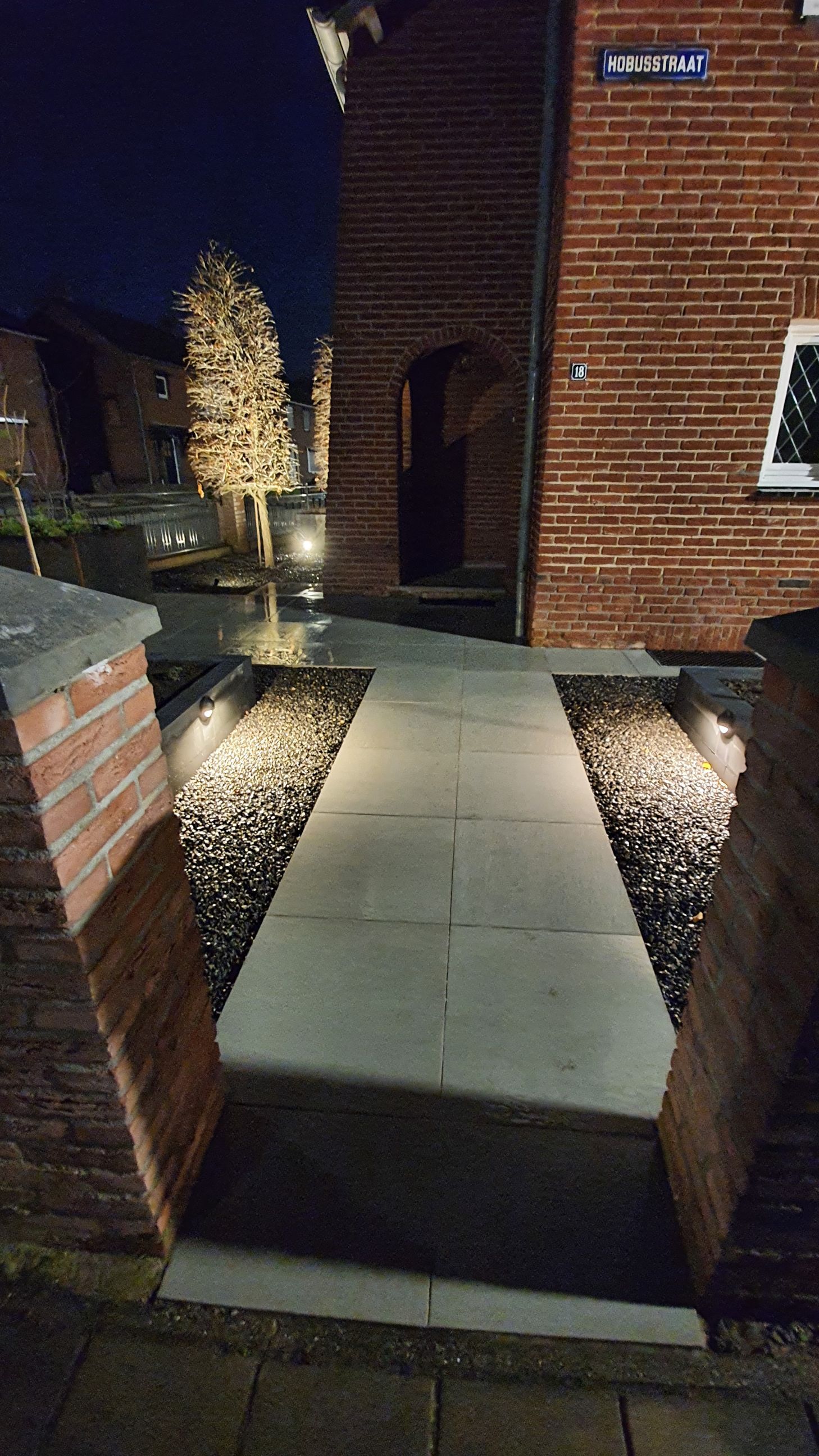 Een smal, verlicht bakstenen pad loopt 's nachts langs een bakstenen gebouw, dat langs een klein beplant gebied naar een deuropening leidt.