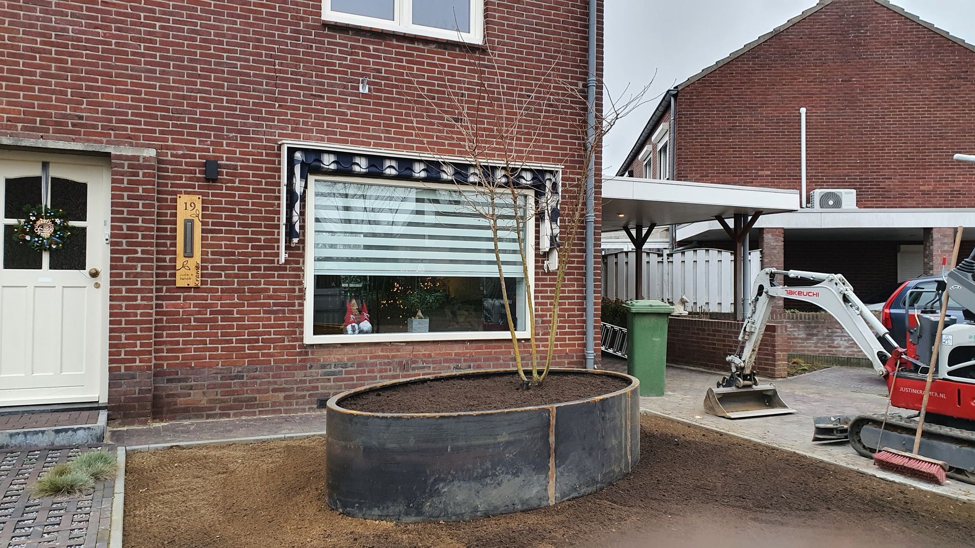 Bakstenen huis met een raam aan de voorzijde, een ronde plantenbak en een kleine carport met gereedschap en apparatuur in de buurt.