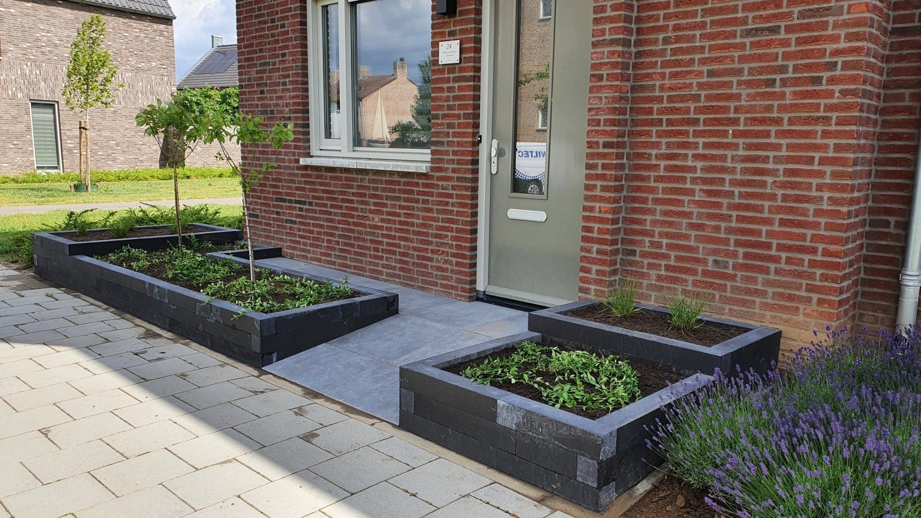 Vooringang met bakstenen muren, glazen deur en twee verhoogde zwarte plantenbakken langs een geplaveid pad.