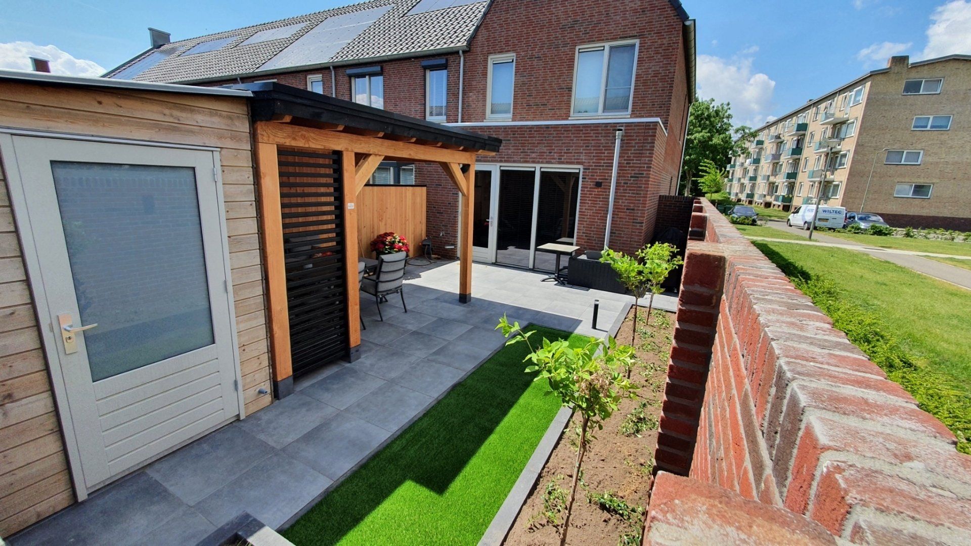 Terras in de achtertuin naast bakstenen huizen, met een klein schuurtje, kunstgras en een laag muurtje langs het pad.
