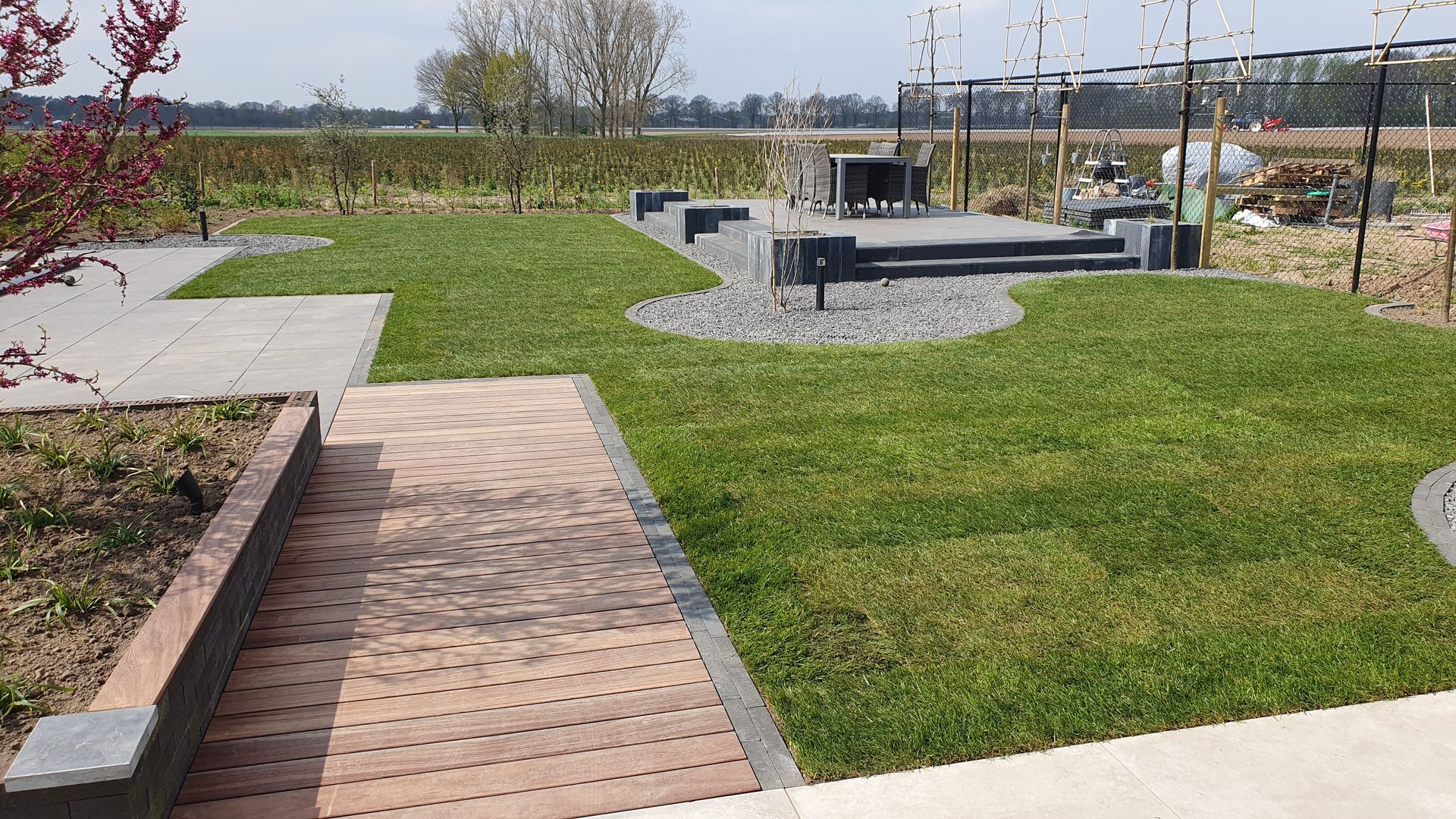 Een bakstenen pad door een aangelegd park met gras, een zitgedeelte met grind en bankjes bij een vijver.