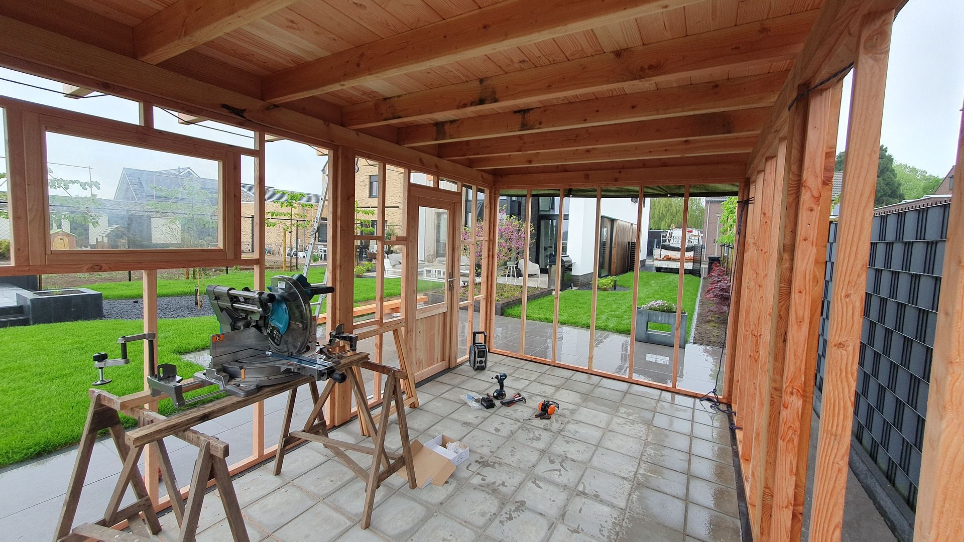 Houten veranda in aanbouw met gereedschap, steigers en stenen vloer naast een grasveld.