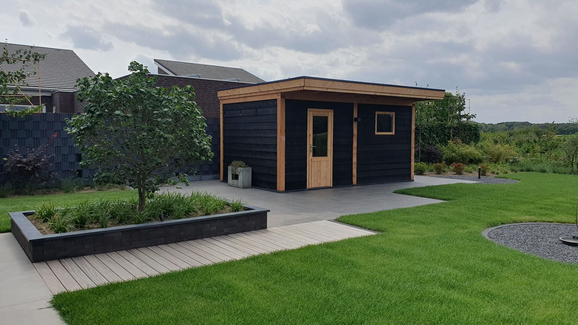 Een klein zwart tuinhuisje met houten afwerking staat naast een gazon en verhoogde plantenbakken onder een bewolkte hemel.