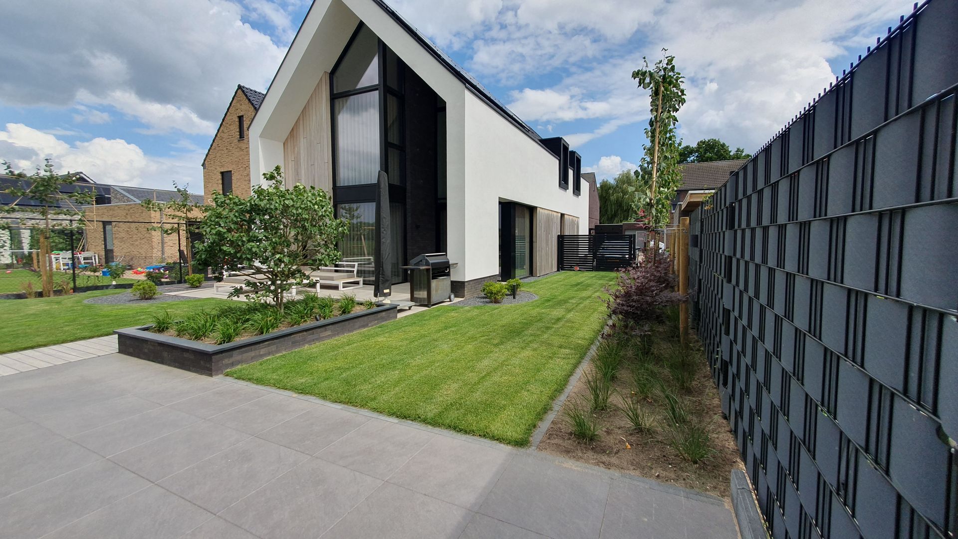 Modern huis met schuin dak, groen gazon, betonnen oprit en een donkere schutting onder een gedeeltelijk bewolkte hemel.