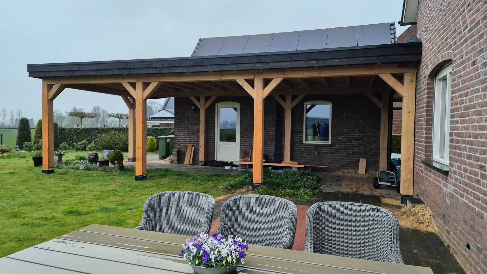 Overdekt terras met houten palen, zonnepanelen op het dak en zitgelegenheid buiten naast een bakstenen huis.
