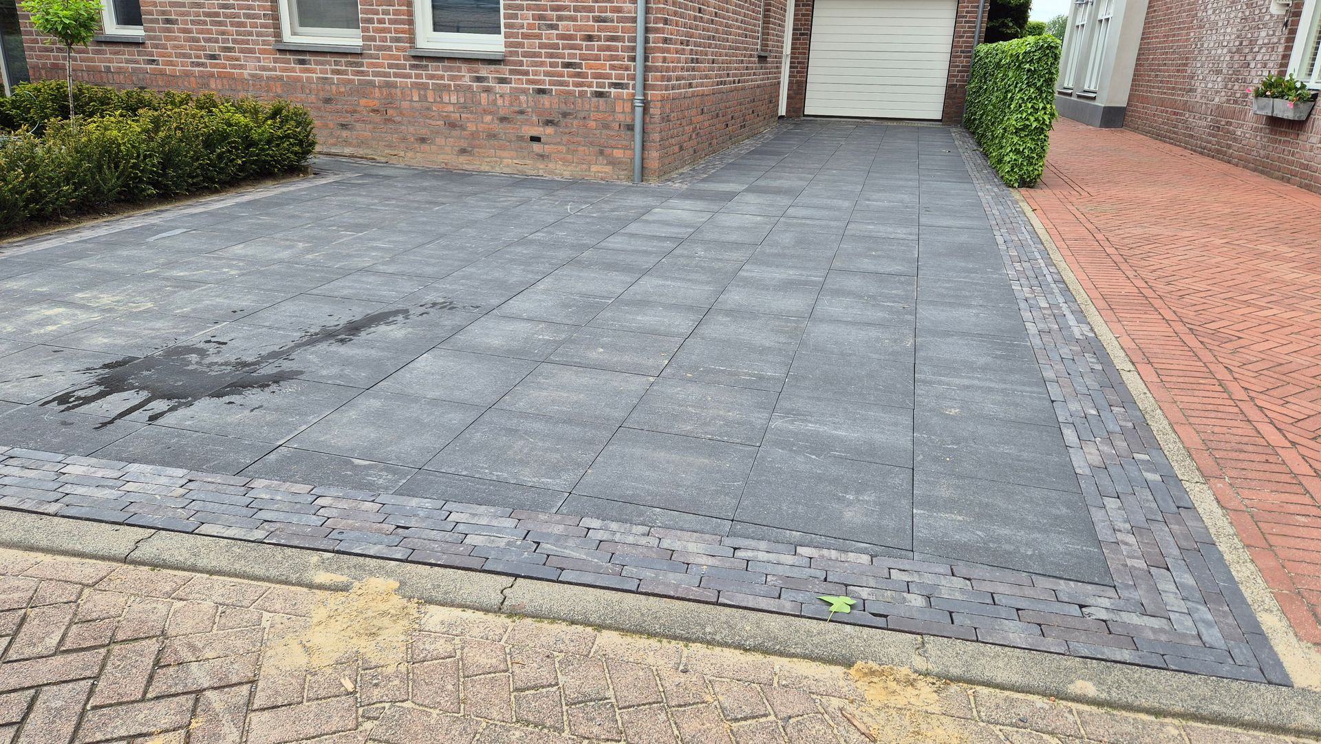 Een geplaveide oprit naast een bakstenen huis, met een donkergrijs terras en een rand van lichtbruine natuursteen.