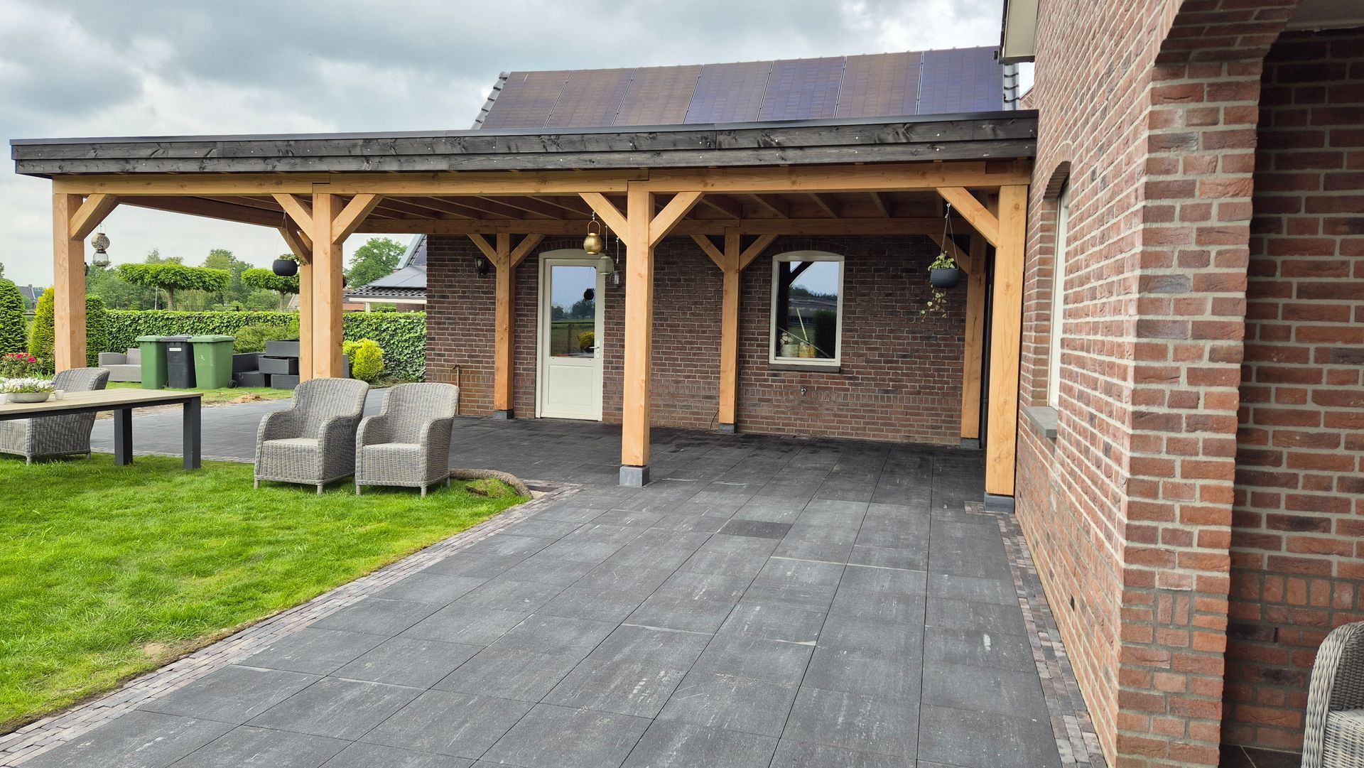 Overdekt terras van baksteen met houten palen, tuinmeubilair en een grasveld daarachter.