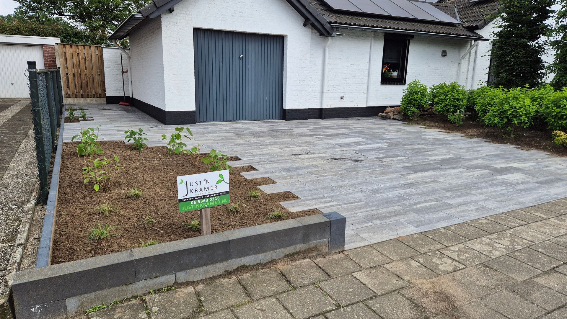 Kleine witte garage met grijze deur, verharde oprit en nieuw aangelegde voortuin.