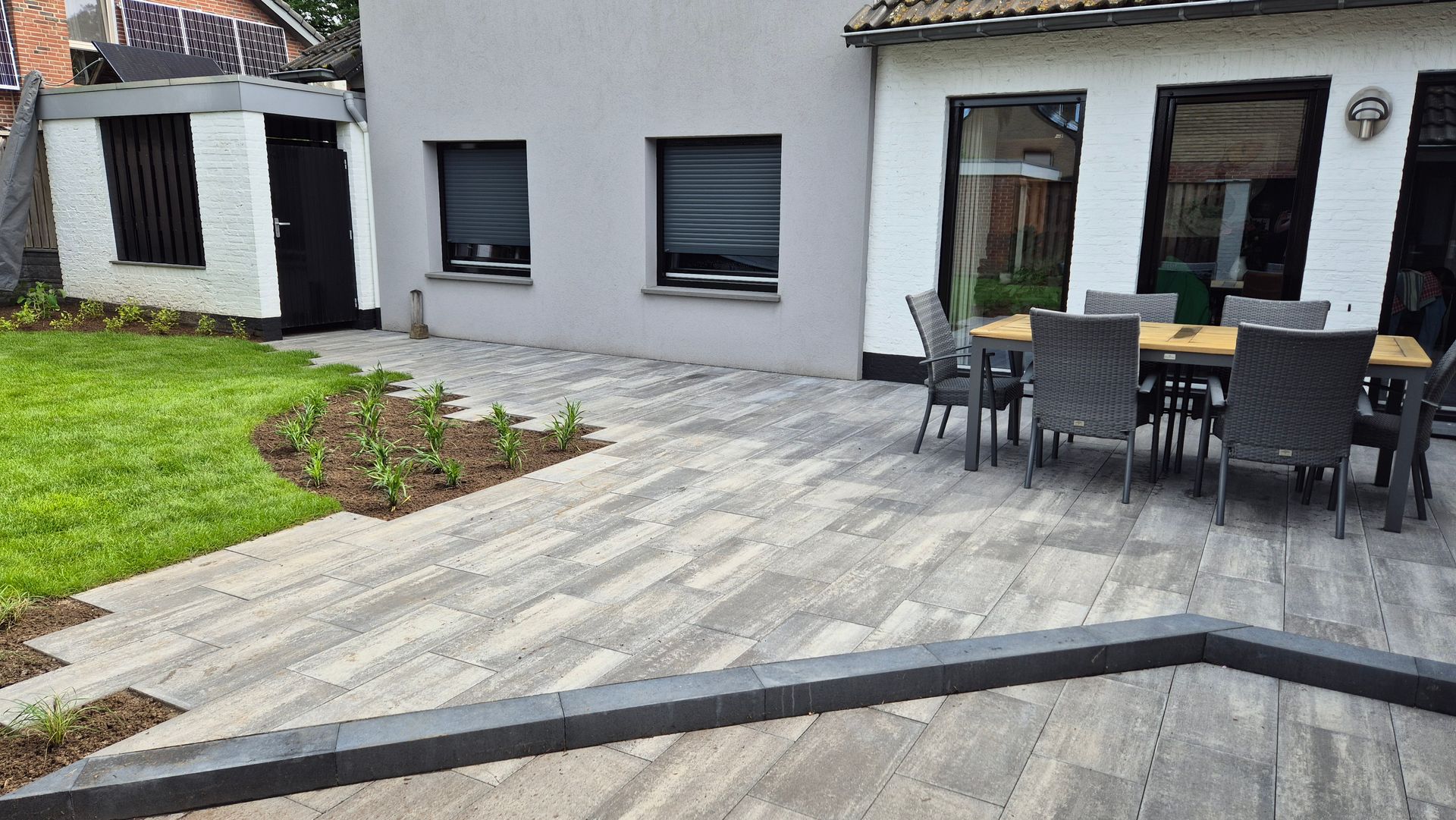 Terras met grijze stenen bestrating, zwarte tuinset en een kleine tuin naast een modern huis.