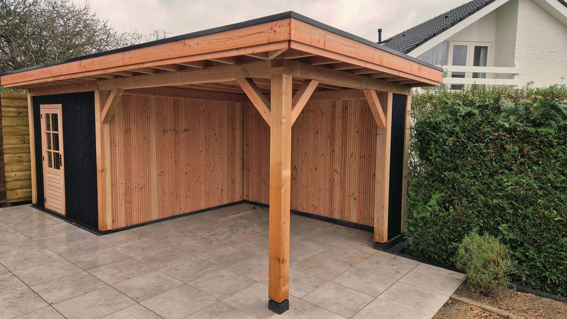 Houten carport of schuur met een schuin dak op een verharde oprit naast een heg en een huis.