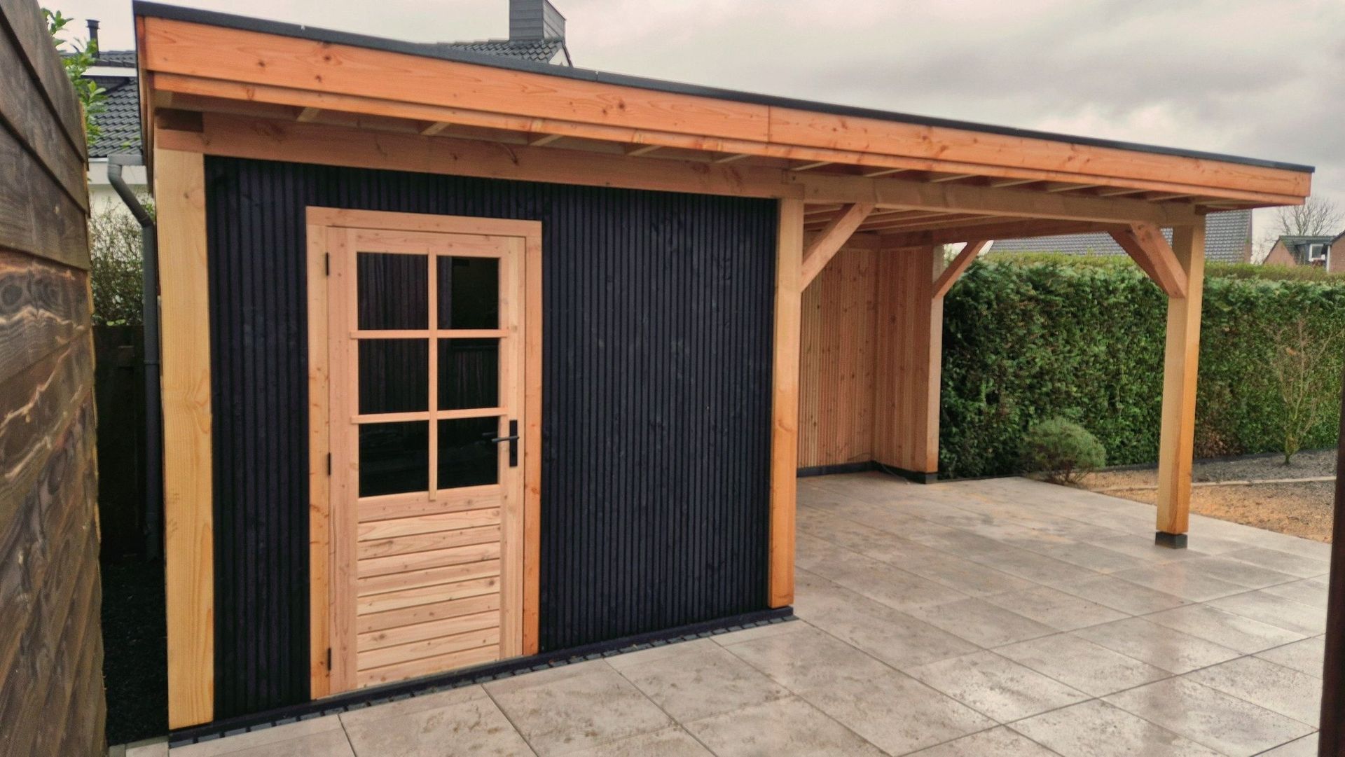 Houten terrasoverkapping met een lichte houten deur en zwarte gevelbekleding naast een betegelde tuin.