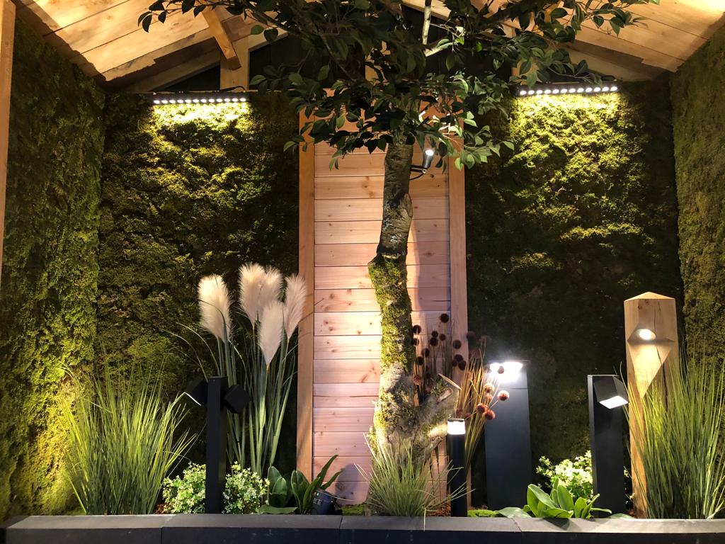 Tuindecoratie met hoog gras, witte pluimen en een centraal, met klimplanten begroeid houten paneel onder warme verlichting.