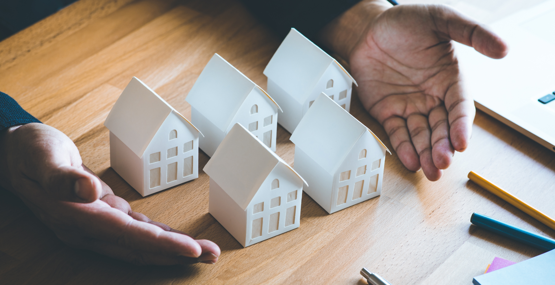 Check Out Our Consulting Services! A person is holding a handful of paper houses on a wooden table.