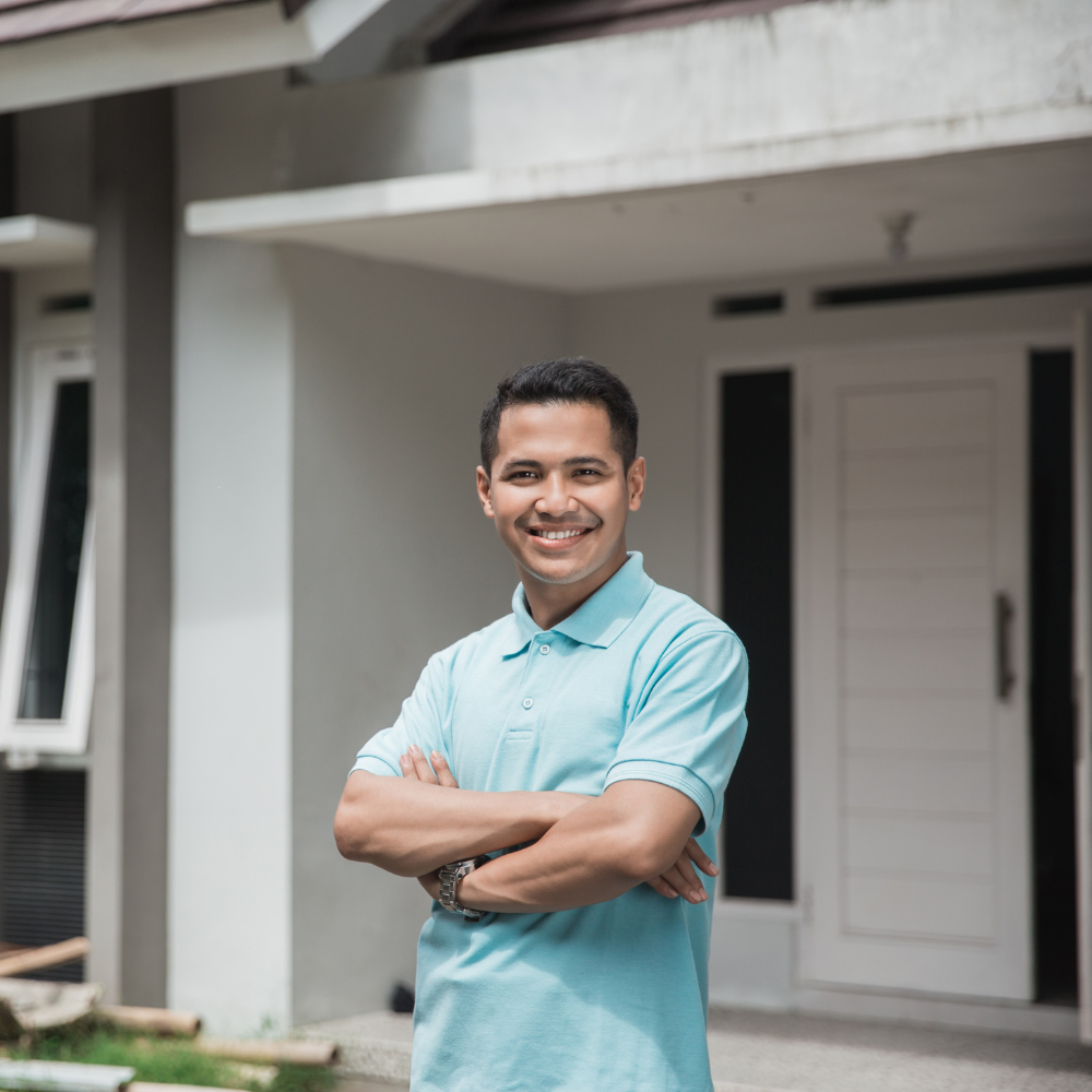 A man in a blue shirt is standing in front of a house with his arms crossed.