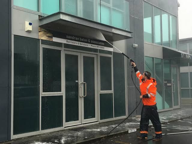 Worker in orange safety gear pressure-washes a glass storefront entrance.