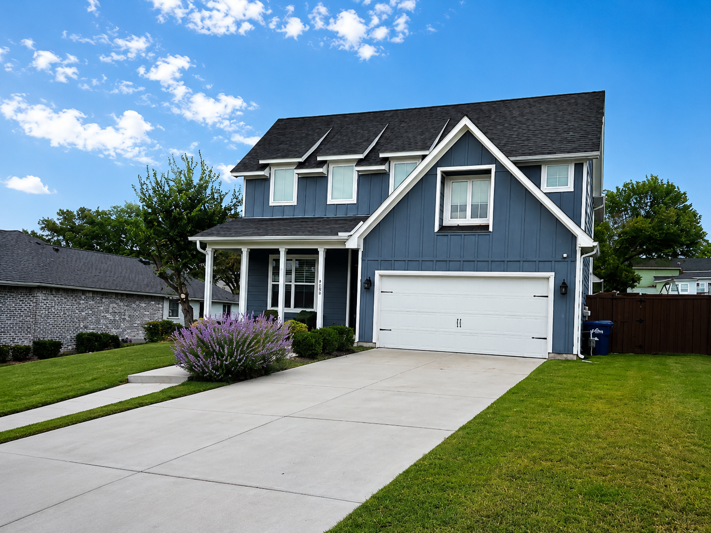 Blue two-story suburban house with white trim, front porch, garage, and a wide driveway under a blue sky